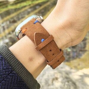 Puede incluir: Una correa de reloj de cuero marr&oacute;n con costuras azules y naranjas est&aacute; sujeta a una mu&ntilde;eca. La esfera del reloj es plateada. La correa tiene un cierre de hebilla. La correa est&aacute; hecha de un material similar al ante.