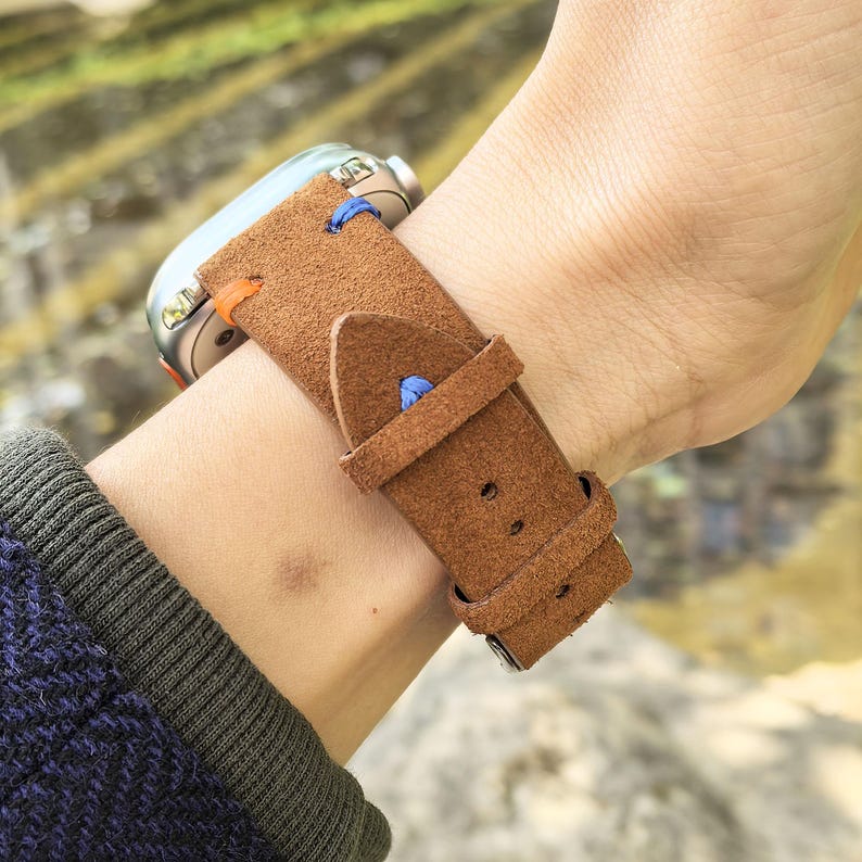 Puede incluir: Una correa de reloj de cuero marr&oacute;n con costuras azules y naranjas est&aacute; sujeta a una mu&ntilde;eca. La esfera del reloj es plateada. La correa tiene un cierre de hebilla. La correa est&aacute; hecha de un material similar al ante.