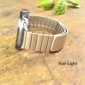 May include: A smartwatch with a dark gray metallic casing and a light beige, segmented metal link band. The watch features a side crown with a red accent ring. The text Star Light is printed in black in the lower right corner of the image.