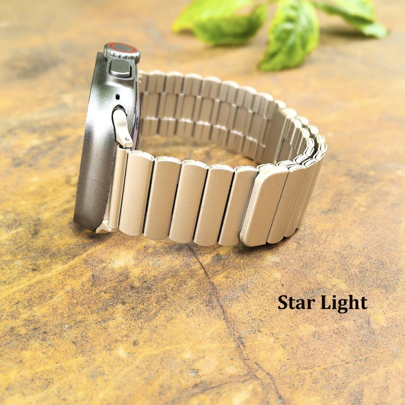 May include: A smartwatch with a dark gray metallic casing and a light beige, segmented metal link band. The watch features a side crown with a red accent ring. The text Star Light is printed in black in the lower right corner of the image.