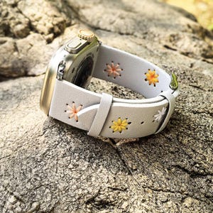 Pode incluir: Pulseira de relógio de couro branco com flores bordadas em tons de amarelo, laranja e rosa. A pulseira é projetada para um Apple Watch.