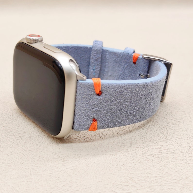 Puede incluir: Un reloj inteligente plateado con pantalla negra y correa de reloj de ante azul claro. La correa tiene costuras naranjas y una hebilla plateada. El reloj se muestra sobre una superficie beige claro.