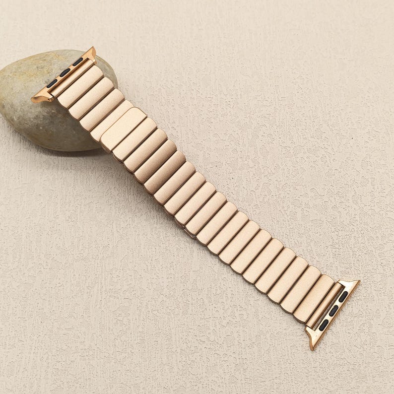 May include: A rose gold-colored metal watch band featuring an expandable, link-style design with a smooth, matte finish. The band includes integrated attachment connectors at both ends, each featuring small black rectangular slots for secure fastening.