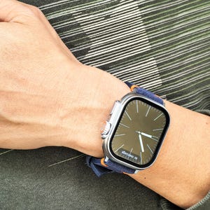Puede incluir: Un reloj inteligente plateado con una pantalla oscura que muestra la hora, usado en una mu&ntilde;eca. El reloj tiene una correa azul marino con detalles en naranja. El fondo muestra un patr&oacute;n de rayas verdes y blancas.
