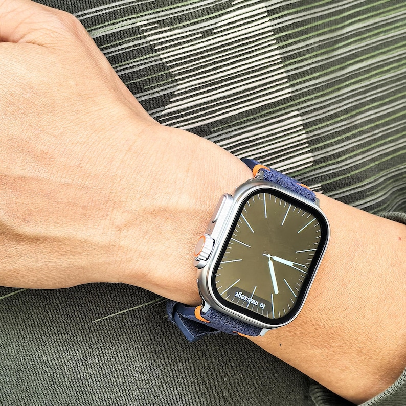 Puede incluir: Un reloj inteligente plateado con una pantalla oscura que muestra la hora, usado en una mu&ntilde;eca. El reloj tiene una correa azul marino con detalles en naranja. El fondo muestra un patr&oacute;n de rayas verdes y blancas.