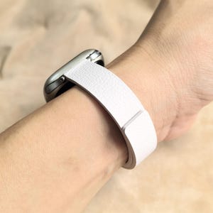 May include: A white leather watch band is wrapped around a wrist, attached to a silver smartwatch. The band has a textured surface and a simple, modern design. The watch is a stylish accessory.