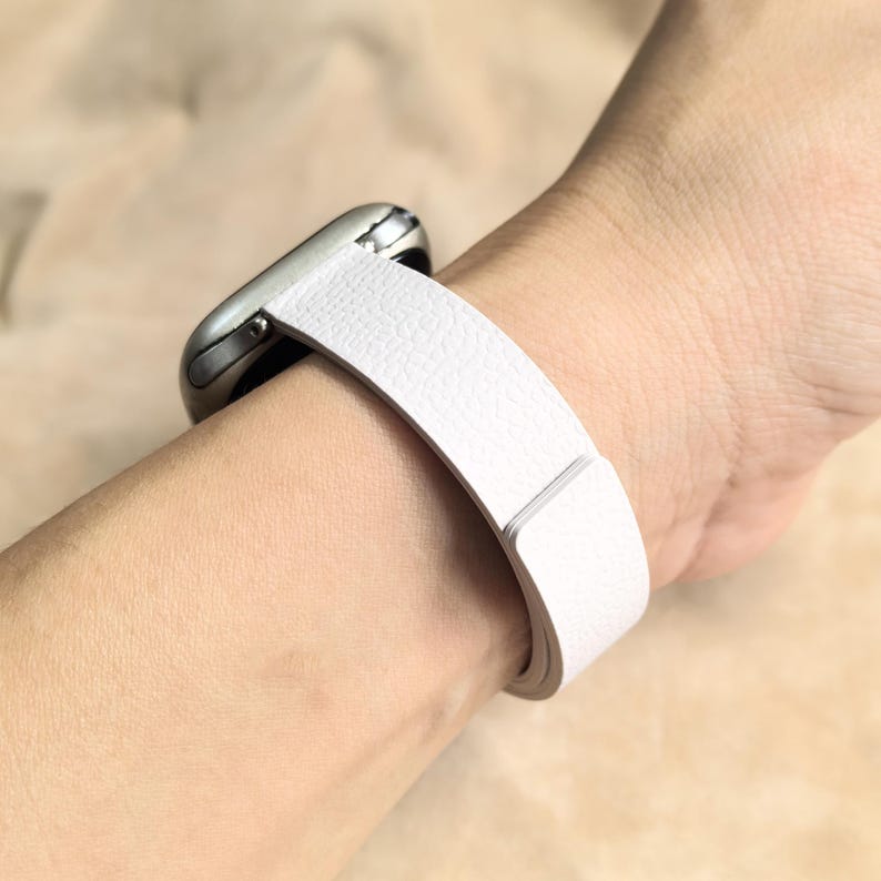 May include: A white leather watch band is wrapped around a wrist, attached to a silver smartwatch. The band has a textured surface and a simple, modern design. The watch is a stylish accessory.