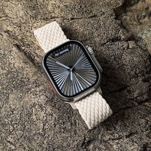 Könnte beinhalten: Eine silberne Apple Watch mit einem weißen geflochtenen Armband. Das Zifferblatt der Uhr zeigt ein radiales Muster mit dem Text "No messag" an.