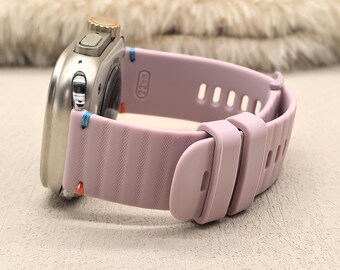 May include: A silver smartwatch with a light purple silicone band. The band has a ribbed texture and is secured with a matching clasp. The watch face is round with a silver bezel and a small orange accent.
