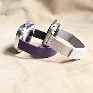 May include: Two smartwatches with silver cases and different colored bands. One watch has a purple band, and the other has a white band. The bands are made of a textured material. The watches are resting on a neutral-colored surface.