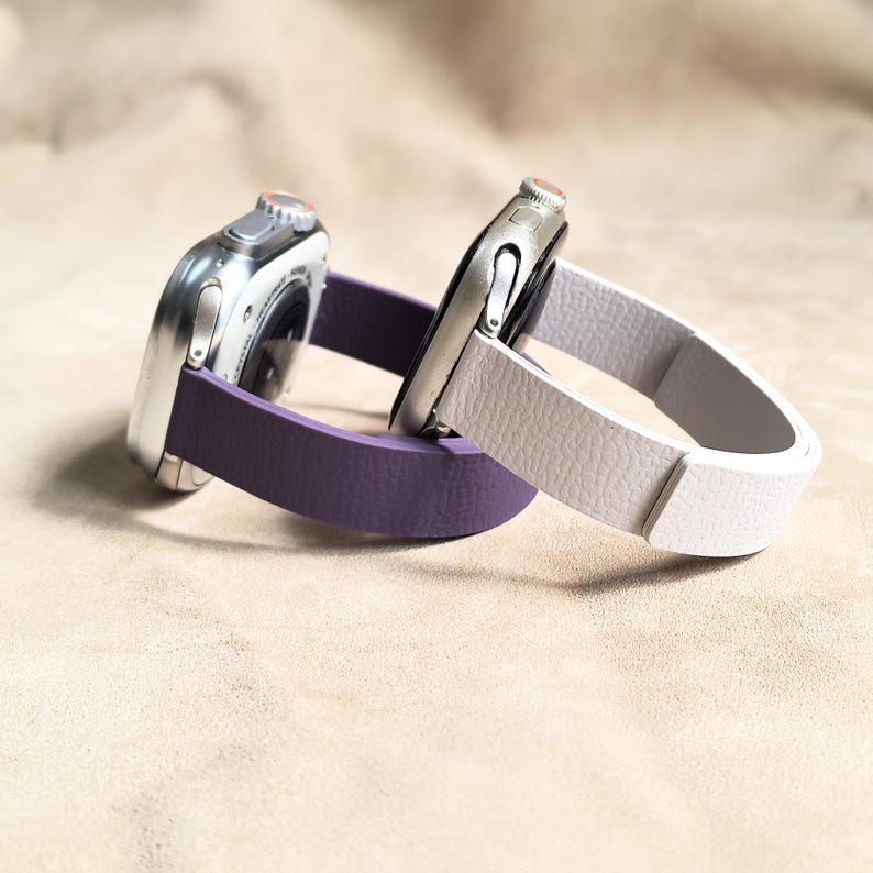 May include: Two smartwatches with silver cases and different colored bands. One watch has a purple band, and the other has a white band. The bands are made of a textured material. The watches are resting on a neutral-colored surface.