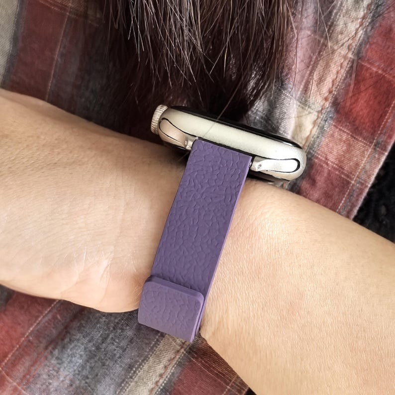 May include: A silver smartwatch with a textured purple band is shown on a wrist. The band is a solid color and appears to be made of leather or a similar material. The watch face is a light color, and the band is secured with a simple clasp.