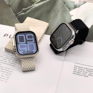 Può includere: Due smartwatch con casse argentate e display digitali. Uno ha un cinturino beige intrecciato, l'altro un cinturino nero intrecciato. Gli schermi mostrano l'ora e un messaggio. Gli orologi sono su una superficie bianca.