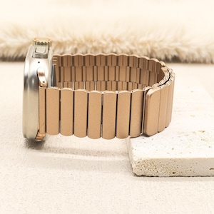 May include: A metallic rose gold watch band with a segmented, flexible link design attached to a silver-toned smartwatch casing. The band features a matte finish and a continuous, expandable structure that connects directly to the watch housing.