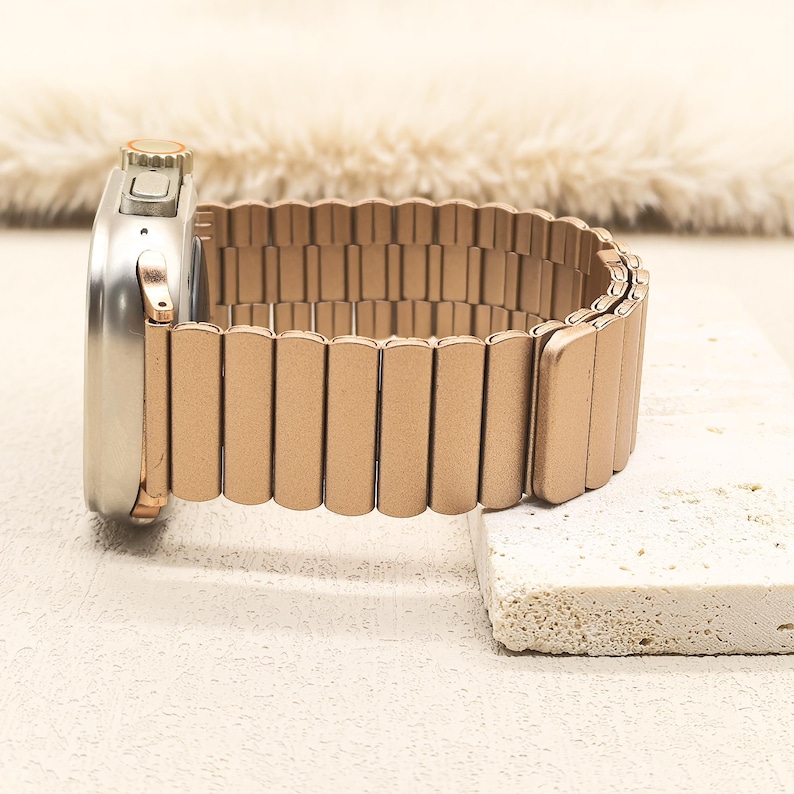 May include: A metallic rose gold watch band with a segmented, flexible link design attached to a silver-toned smartwatch casing. The band features a matte finish and a continuous, expandable structure that connects directly to the watch housing.