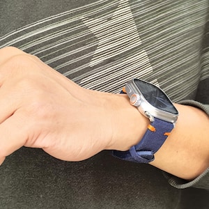 Puede incluir: Un reloj inteligente plateado con pantalla oscura y detalles en naranja se lleva en la mu&ntilde;eca. El reloj tiene una correa azul marino con costuras naranjas. La persona lleva una camisa de manga larga verde oscuro.