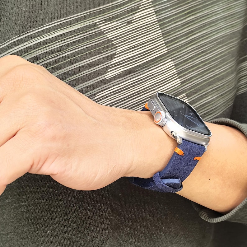 Puede incluir: Un reloj inteligente plateado con pantalla oscura y detalles en naranja se lleva en la mu&ntilde;eca. El reloj tiene una correa azul marino con costuras naranjas. La persona lleva una camisa de manga larga verde oscuro.
