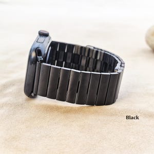 May include: A black smartwatch with a matching black metal link band. The watch features a rectangular case with a side button and a digital crown. The band consists of multiple rectangular segments connected together in a continuous loop design.