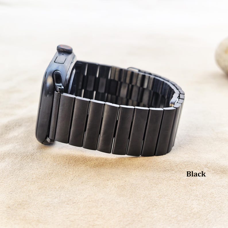 May include: A black smartwatch with a matching black metal link band. The watch features a rectangular case with a side button and a digital crown. The band consists of multiple rectangular segments connected together in a continuous loop design.