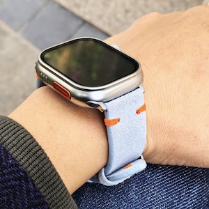 Puede incluir: Un reloj inteligente plateado con pantalla negra y correa de ante azul claro con costuras naranjas. El reloj est&aacute; en una mu&ntilde;eca, mostrando el dise&ntilde;o del reloj y la correa.