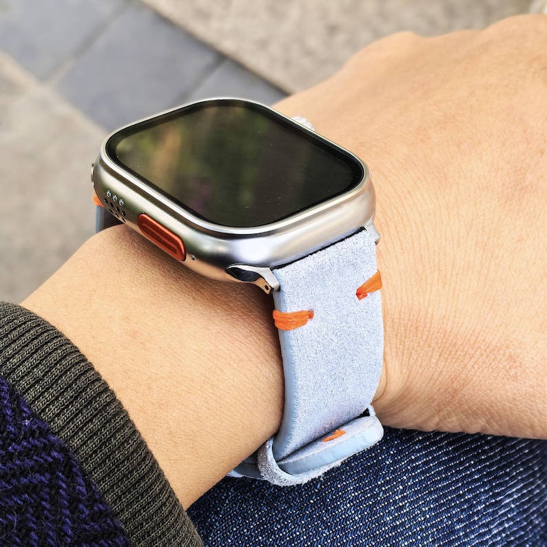 Puede incluir: Un reloj inteligente plateado con pantalla negra y correa de ante azul claro con costuras naranjas. El reloj est&aacute; en una mu&ntilde;eca, mostrando el dise&ntilde;o del reloj y la correa.