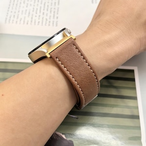 May include: A wristwatch featuring a textured brown leather strap with white contrast stitching. The watch case is constructed from a combination of silver-toned metal and gold-toned accents, showcasing a circular design with a side-mounted button.