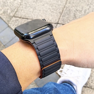 Op de afbeelding: Zwarte smartwatch met een zwart bandje met een rechthoekig schakelontwerp en oranje accenten. Het horloge wordt om een pols gedragen. Op de achtergrond zijn een blauwe mouw en een witte schoen te zien.