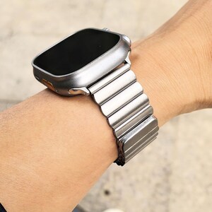 以下が含まれることがあります： 銀色のApple Watchと銀色のメタルバンド。時計の文字盤は黒で、バンドは小さな連結されたリンクのシリーズで構成されています。