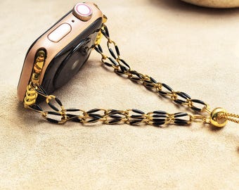 May include: A gold-tone smartwatch with a black screen and a chain-link band. The band features alternating gold and black oval links. The watch is resting on a light tan surface.
