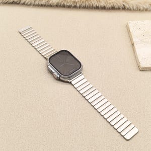 May include: A silver-toned smartwatch with a rectangular face and a dark, sunburst-patterned display. The watch features a metallic link band with rectangular segments and a side crown with a red accent, presenting a sleek and uniform appearance.