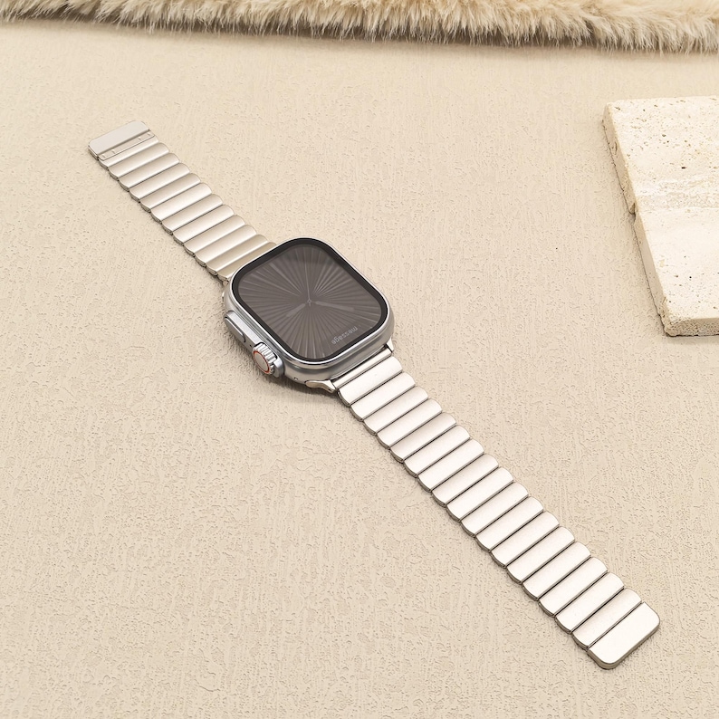 May include: A silver-toned smartwatch with a rectangular face and a dark, sunburst-patterned display. The watch features a metallic link band with rectangular segments and a side crown with a red accent, presenting a sleek and uniform appearance.