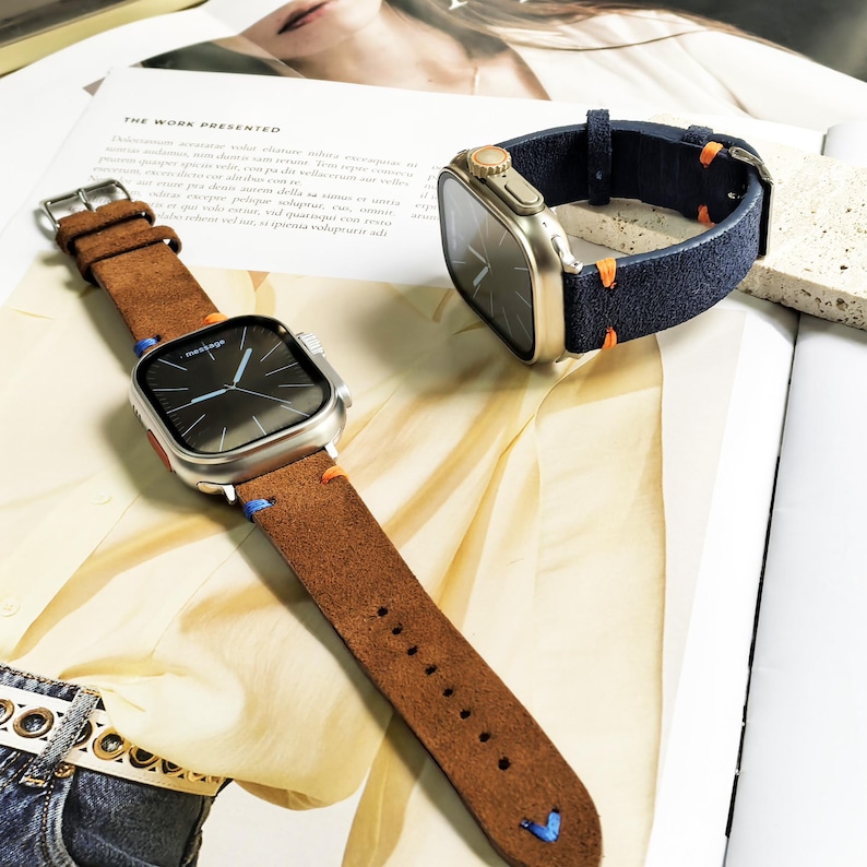Puede incluir: Dos smartwatches con correas de cuero se muestran en una revista. Uno tiene una correa de cuero marr&oacute;n con costuras azules y naranjas. El otro tiene una correa azul marino con costuras naranjas. Las esferas de los relojes son negras.