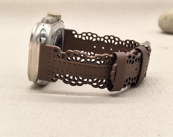 May include: A silver smartwatch with a brown leather strap. The strap has a decorative, openwork design. The watch is displayed on a neutral surface, highlighting the details of the watch and strap.