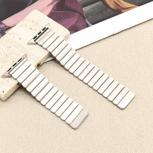 May include: A silver-toned metal watch band featuring a series of rectangular, interlocking links. The band is designed with a flexible, expandable construction and includes integrated attachment connectors at both ends for a secure watch fit.