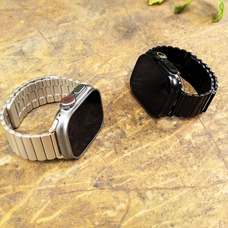 May include: Two smartwatches with rectangular screens and metal link bands. One watch has a silver-toned case and band with a red accent on the side dial, while the other watch features a black case and matching black metal link band.
