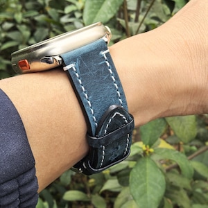 Pode incluir: Um close-up de um smartwatch com uma pulseira de couro azul. A pulseira é costurada com linha branca e tem uma fivela preta. A caixa do relógio é prateada com um botão laranja.