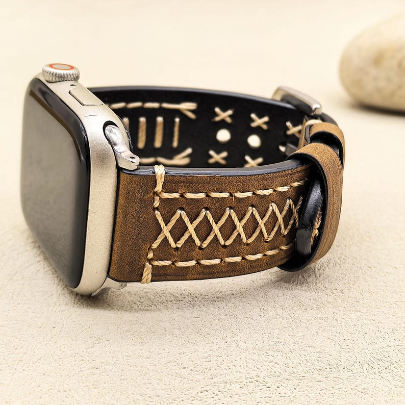 Op de afbeelding: Bruin leren Apple Watch band met witte stiksels en een zwarte gesp. Het band heeft een decoratief patroon van X'en en I's.