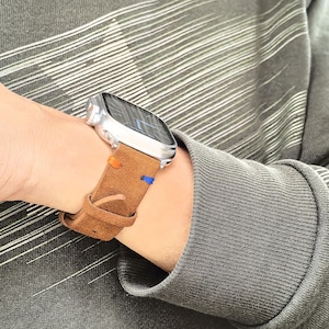 Puede incluir: Un reloj inteligente plateado con una correa de cuero marr&oacute;n. La correa tiene costuras naranjas y azules. El reloj est&aacute; en una mu&ntilde;eca y la persona lleva una camisa gris de manga larga.