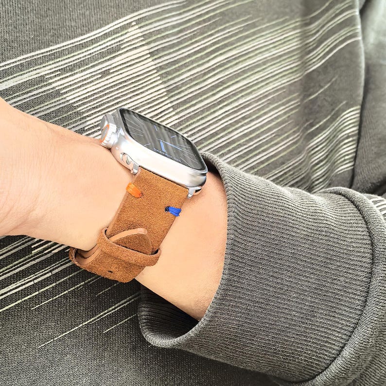 Puede incluir: Un reloj inteligente plateado con una correa de cuero marr&oacute;n. La correa tiene costuras naranjas y azules. El reloj est&aacute; en una mu&ntilde;eca y la persona lleva una camisa gris de manga larga.