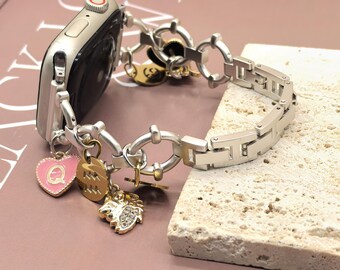 May include: Silver Apple Watch with a decorative bracelet. The bracelet features a chain-link design with various charms, including a pink heart with a letter "Q", a gold charm with a zodiac symbol, and a gold charm with a lion design.