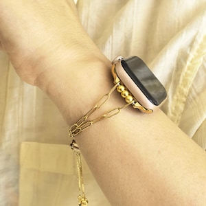 以下が含まれることがあります： ゴールドトーンのApple Watchと、お揃いのチェーンリンクバンド。 バンドは一連の楕円形のリンクと、文字盤の近くにビーズのセクションを備えています。 小さな金の球体が付いた繊細なチェーンがバンドからぶら下がっています。