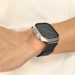 Op de afbeelding: Een zilveren smartwatch met een zwart scherm waarop de tijd en het woord "message" worden weergegeven. Het horloge heeft een zwarte mesh band en een oranje accent. Het horloge zit om een pols.