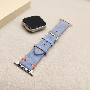 Puede incluir: Correa de reloj de ante azul claro con costuras naranjas y hebilla plateada. La correa se muestra con una esfera de reloj plateada. La correa est&aacute; dise&ntilde;ada para un reloj inteligente.