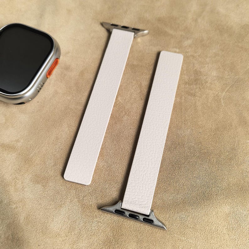 May include: A pair of off-white watch bands with silver connectors, alongside a silver and black smartwatch. The bands have a textured surface and are designed for a smartwatch. The watch and bands are displayed on a tan surface.