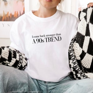 Op de afbeelding: Een wit T-shirt met de tekst "I come back stronger than A 90s TREND" gedrukt op de voorkant. Het model draagt het T-shirt met een blauwe jeans en een zwart-witte cardigan.