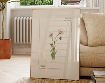 May include: A framed botanical print featuring a watercolor illustration of three pink and white daisies with green stems and leaves. The text "but as she was featuring" and "it felt like breathing" is printed below the illustration. The print is hanging on a wall in a living room setting.