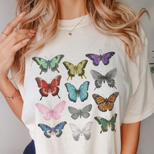 May include: Cream-colored t-shirt featuring a colorful butterfly design. The butterflies are in various colors, including green, yellow, red, blue, pink, and orange. The butterflies are arranged in a grid pattern across the front of the shirt.