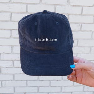 May include: Navy blue corduroy baseball cap with the phrase "i hate it here" embroidered in white. The cap has a curved brim and a button on top. The hat is held up by a hand with blue nail polish.