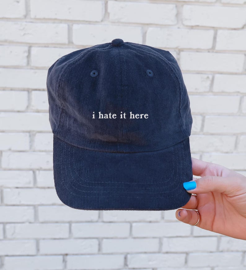 May include: Navy blue corduroy baseball cap with the phrase "i hate it here" embroidered in white. The cap has a curved brim and a button on top. The hat is held up by a hand with blue nail polish.