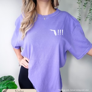 Peut inclure: Un t-shirt violet avec un contour blanc de l'&eacute;tat de Floride et trois points d'exclamation. Le texte "!!!" est sur le t-shirt.